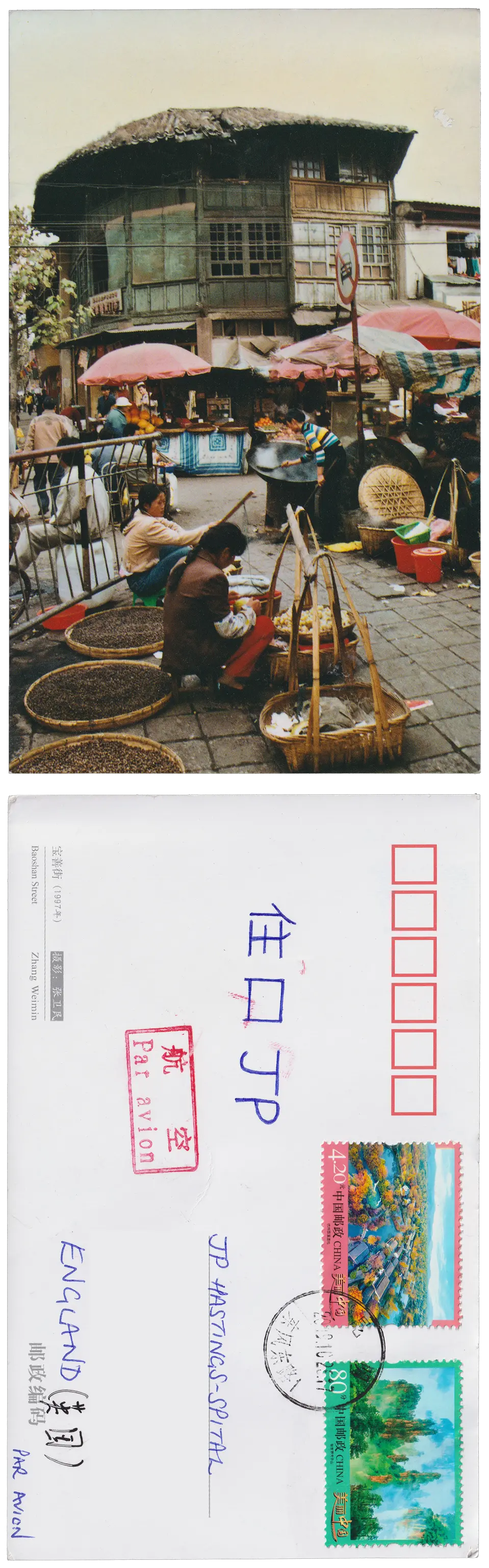 Both sides of a postcard. On the front: A street market in China. Two women sit selling ingredients in the foreground. Red umbrellas cover a stall with a giant cooking wok and selling oranges just beyond. An old, green, two story corner building stands just beyond. On the back: 住口JP