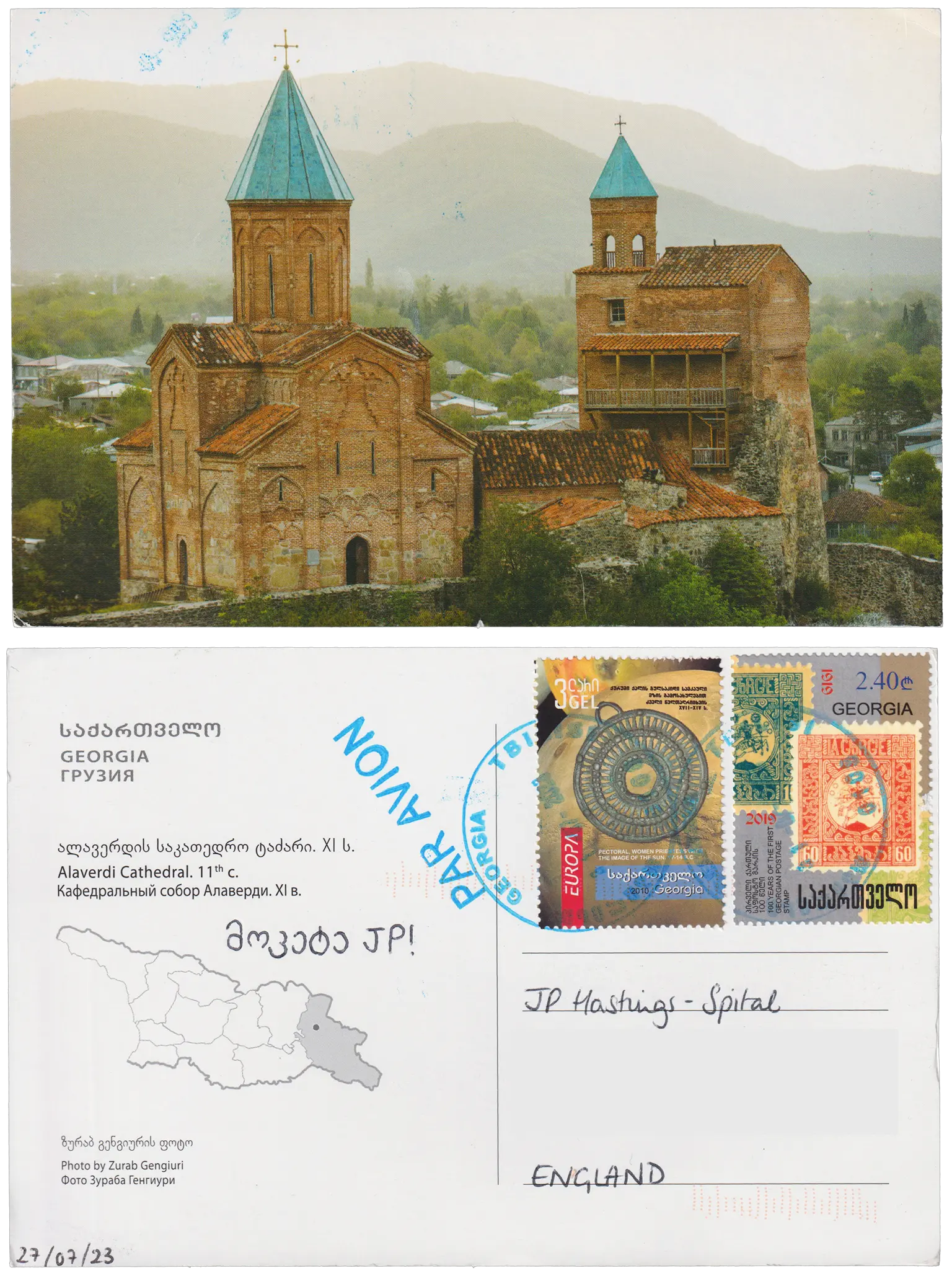 Both sides of a postcard. On the front: A photograph of a large red bricked cathedral with a tall bell tower and a smaller bell tower to the right. The cathedral is surrounded by trees and mountains. On the back: მოკეტე JP!